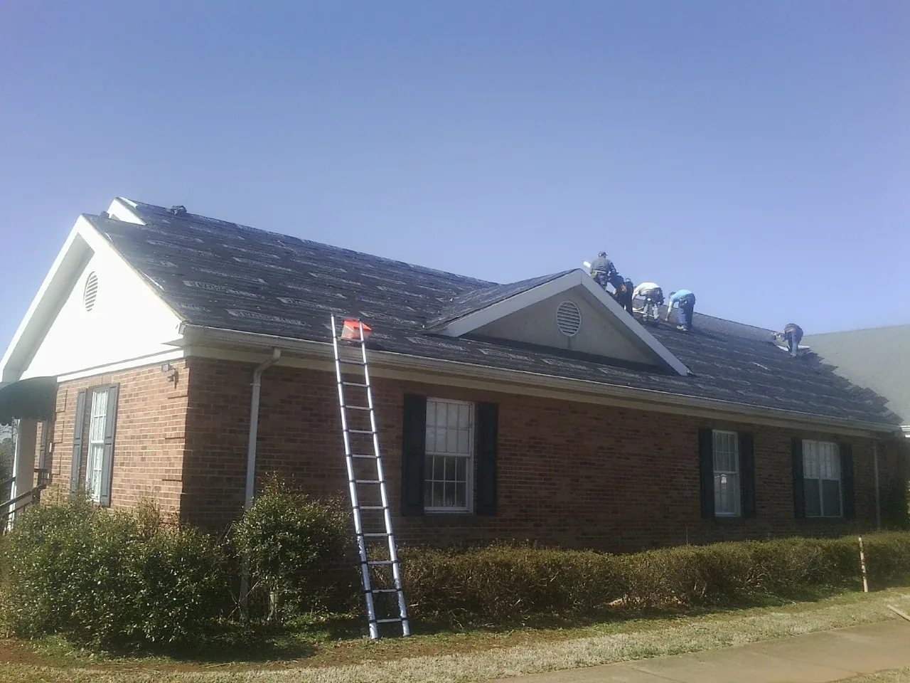 Crew tearing shingles off a brick ranch roof with a ladder set — Metal Roof Repair work by our crew in Schuyler